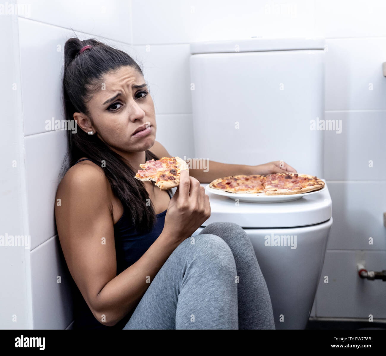Attractive young and sad bulimic young woman feeling guilty and sick eating while sitting on the floor next to the toilet in eating disorders anorexia Stock Photo