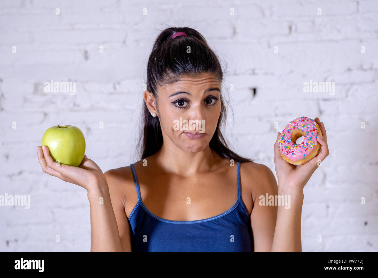 Beautiful young woman tempted having to make choice between apple and doughnut in healthy unhealthy food, detox eating, calories and diet concept. Stock Photo