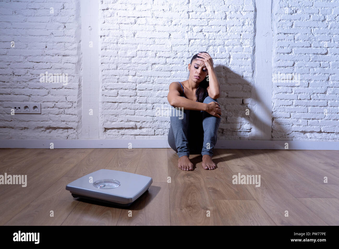 Young anorexic teenager woman sitting alone on ground looking at the scale worried and depressed in dieting and eating disorder concept Stock Photo