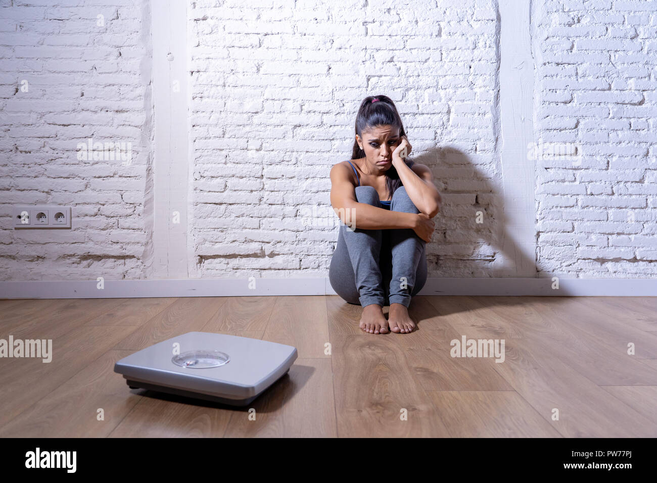Young anorexic bulimic teenager woman sitting alone on ground looking at the scale worried and depressed in failing dieting and eating nutrition disor Stock Photo
