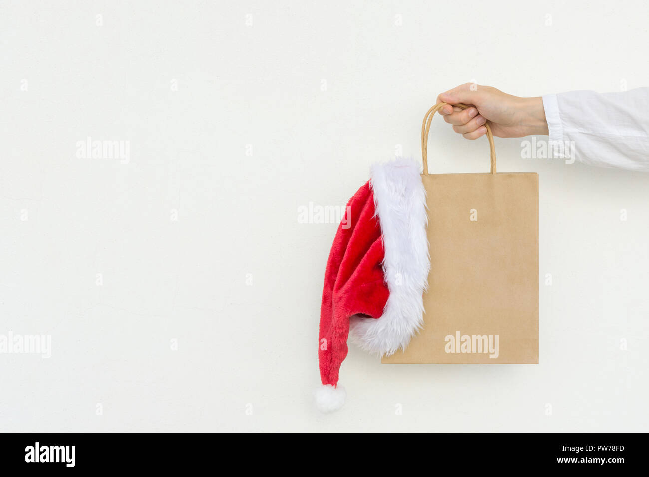 Young caucasian woman holds in hand empty blank mock up brown craft paper bag with hanging santa claus hat on white wall background. Christmas New Yea Stock Photo