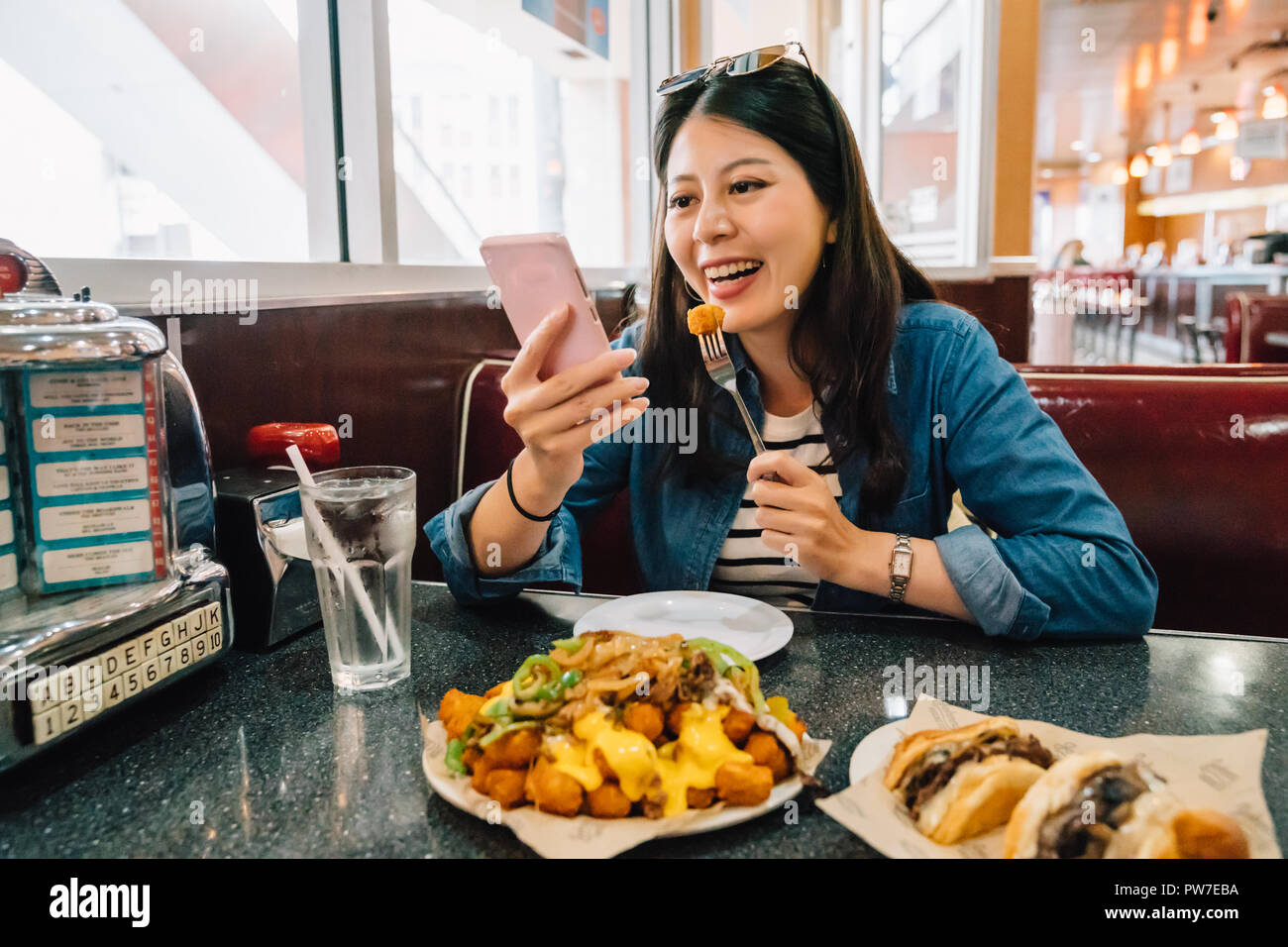 beautiful woman relaxing having lunch in the diner and watching some funny videos by smartphone while eating food. young lady playing games on mobile  Stock Photo