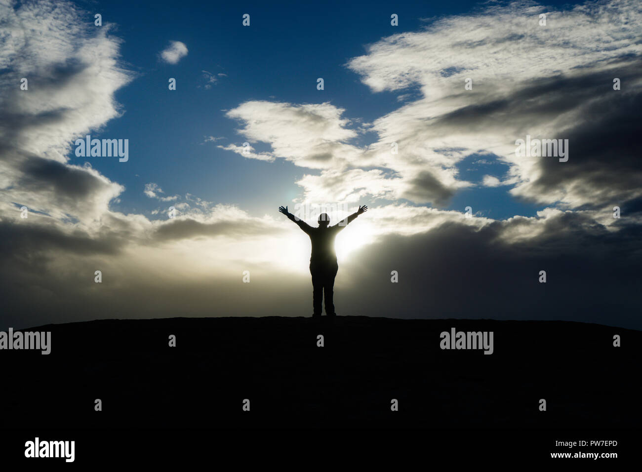 Silhouette of person standing with arms out as sun breaks through clouds. Stock Photo