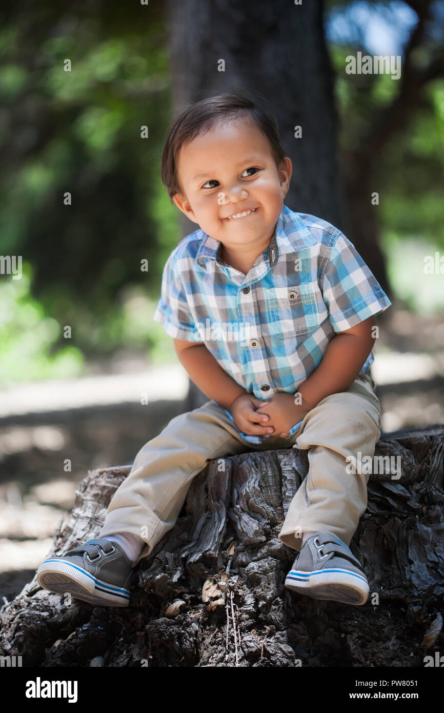 Latino young boy biting his lip with a cute smile, sitting on a tree stump in the outdoors grasping his hands with plaid shirt Stock Photo