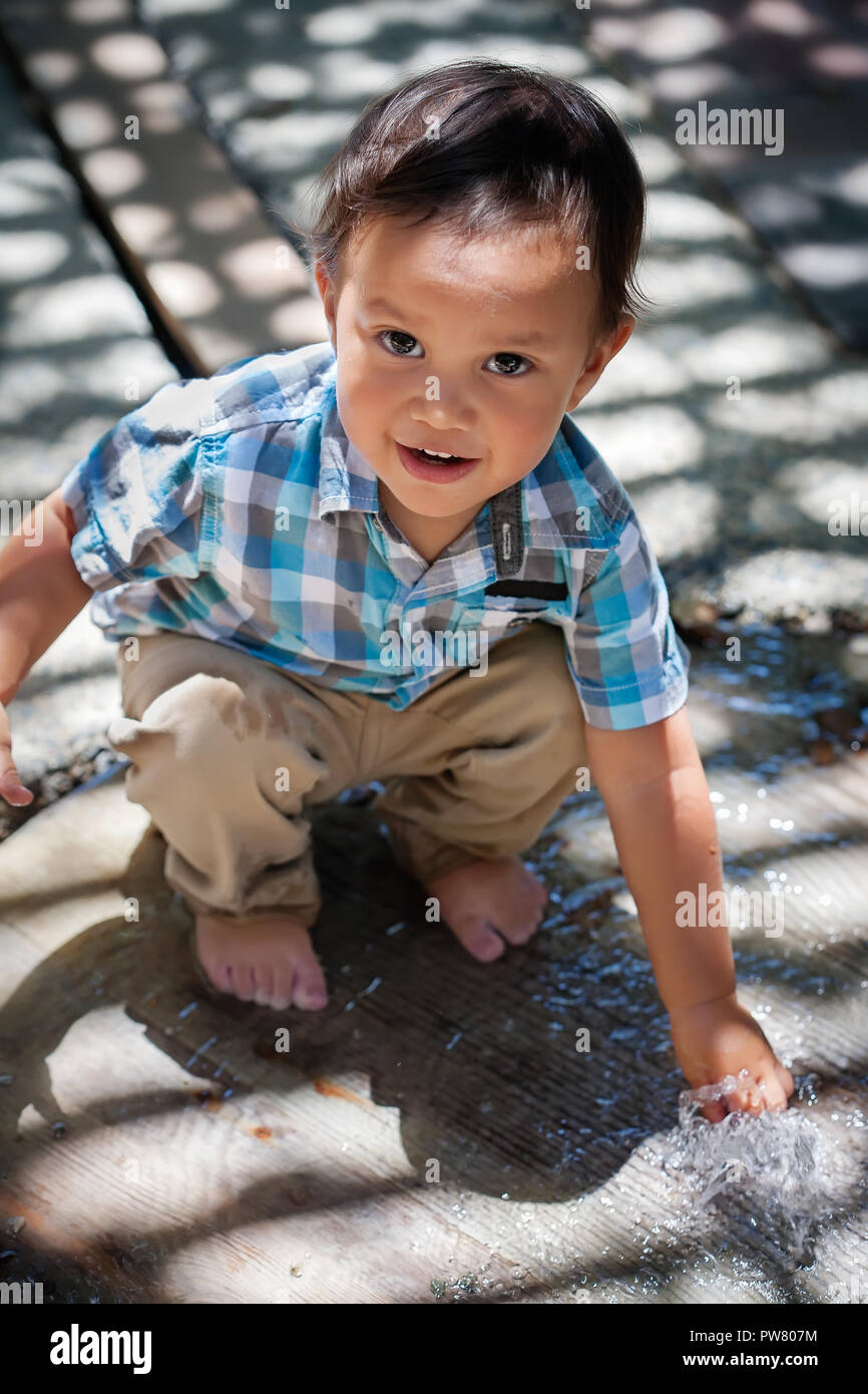 2 year old boy crouching down and touching a water fountain with his hand, getting wet and having fun in summer Stock Photo