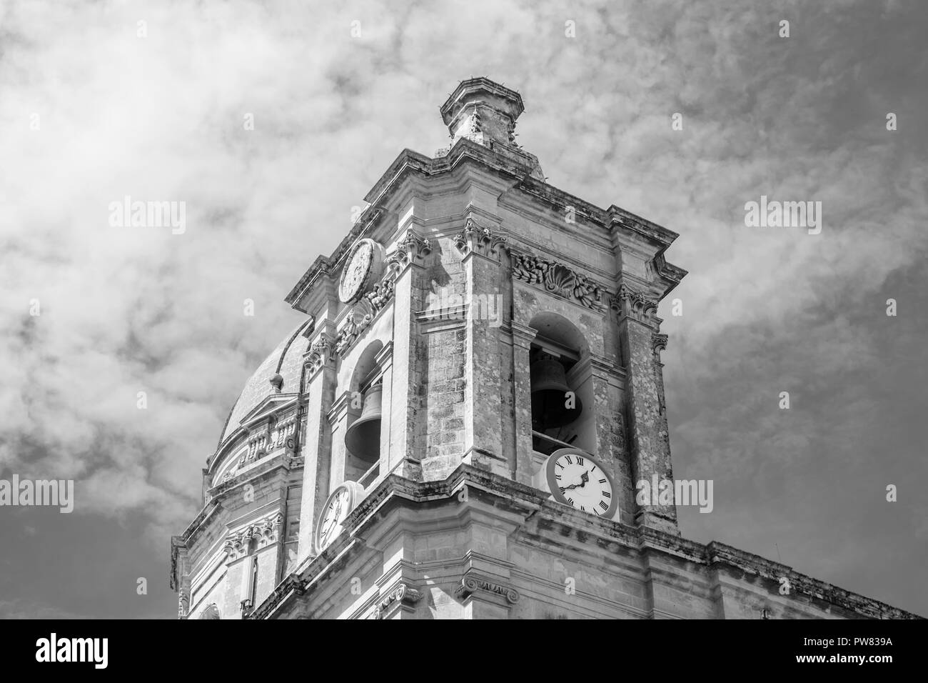 A prominent Old Baroque Belfry of St. Nicholas Church in Siggiewi, Malta in black and white featuring the bells, clocks and a dramatic sky. Stock Photo