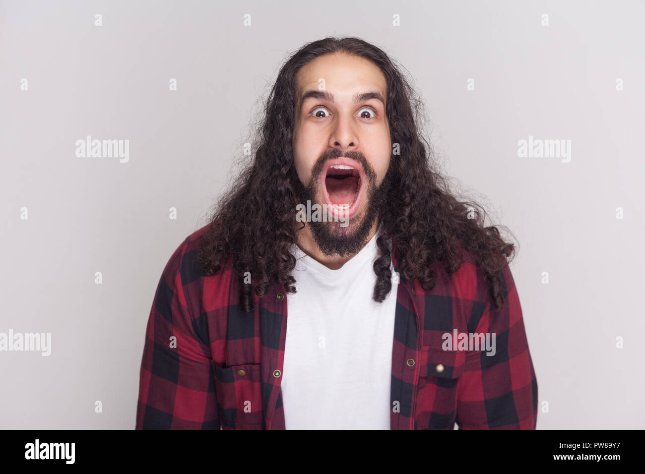 Portrait of amazed handsome man with beard and black long curly hair in casual checkered red shirt looking at camera with open mouth and big eyes. ind Stock Photo