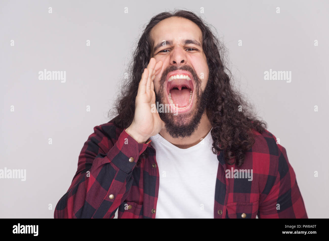 Portrait of roar handsome man with beard and black long curly hair in casual style, checkered red shirt standing and screaming with big open mouth. in Stock Photo