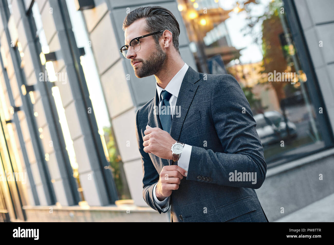 Close up profile portrait of a successful young bearded guy in suit and glasses. So stylish and nerdy. Outdoors on a sunny street, fixing his cuffs Stock Photo