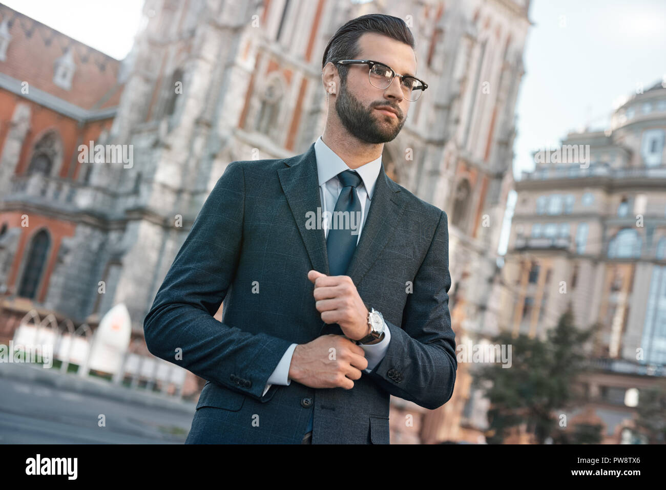 Close up profile portrait of a successful young bearded guy in suit and glasses. So stylish and nerdy. Outdoors on a sunny street, fixing his cuffs Stock Photo