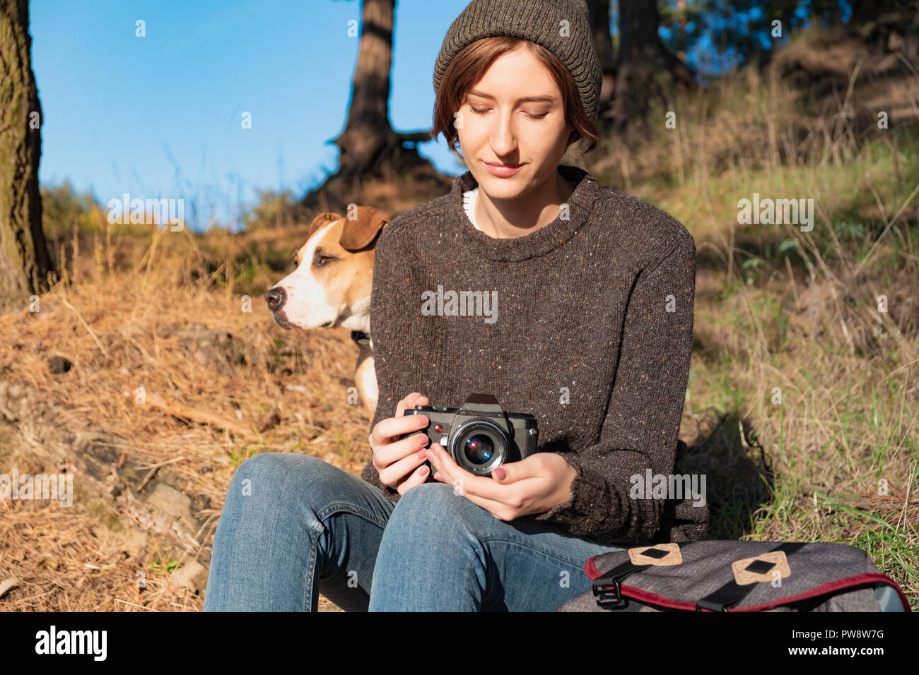 Woman with dog holding a film camera on fine autumn day. Young female hiker sits under pine trees on a sunny afternoon and enjoys fine weather Stock Photo