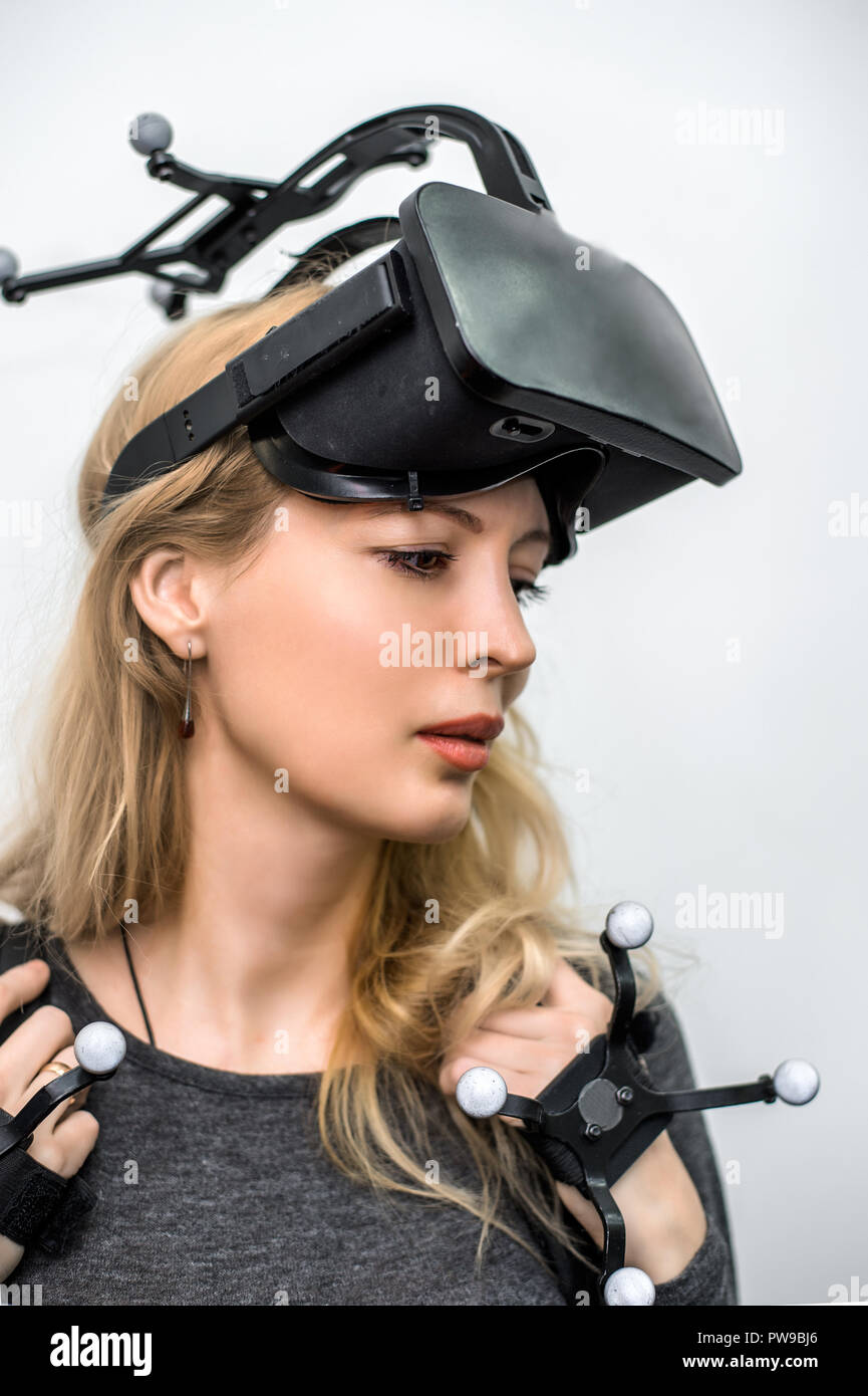 Beautiful woman on grey background. She wearing Virtual reality glasses or goggles on forehead, processor in backpack on back and sensors for tracking Stock Photo