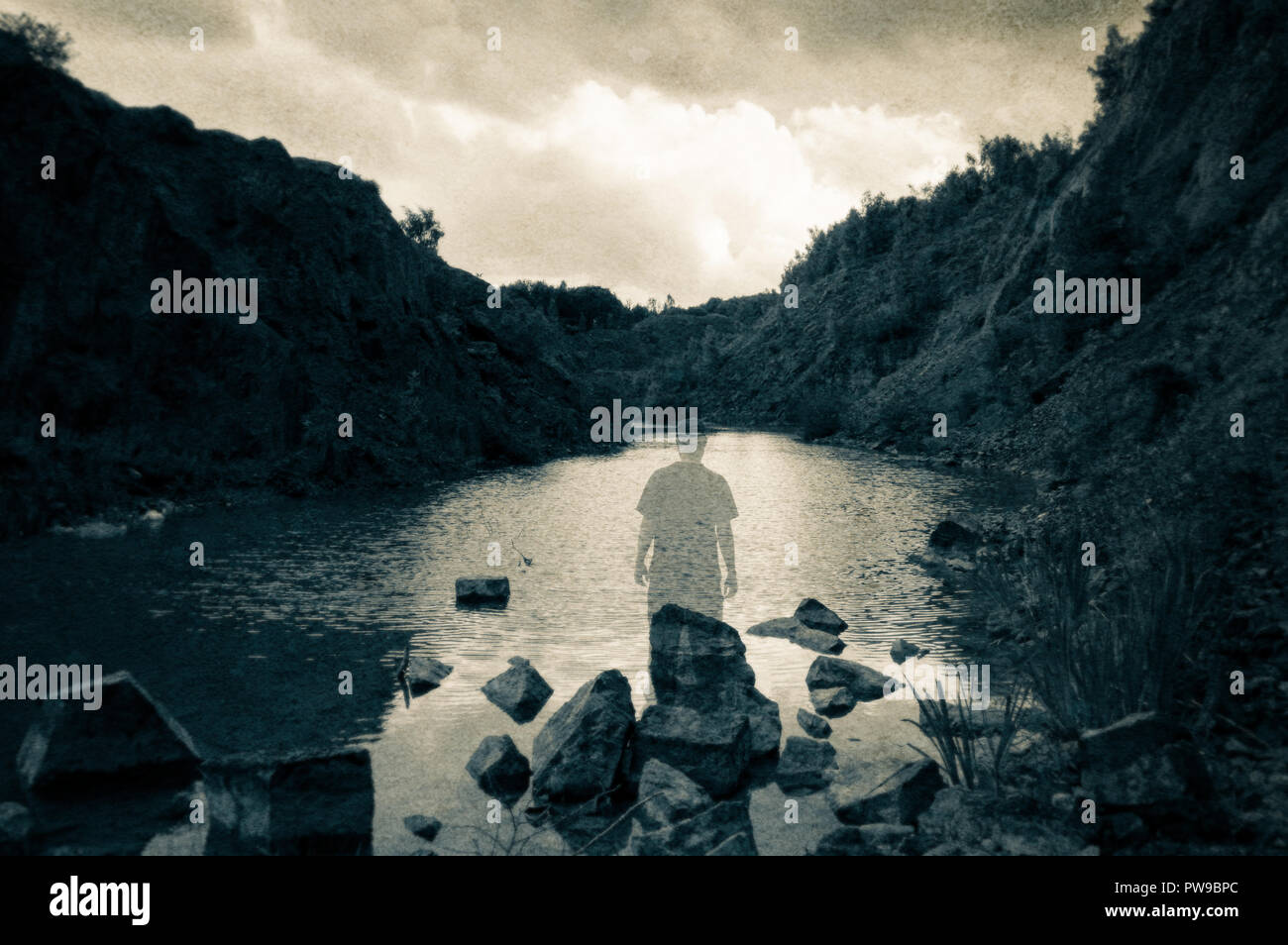 A ghostly transparent figure standing on rocks looking out across a quarry lake surrounded by cliffs. With a muted edit Stock Photo