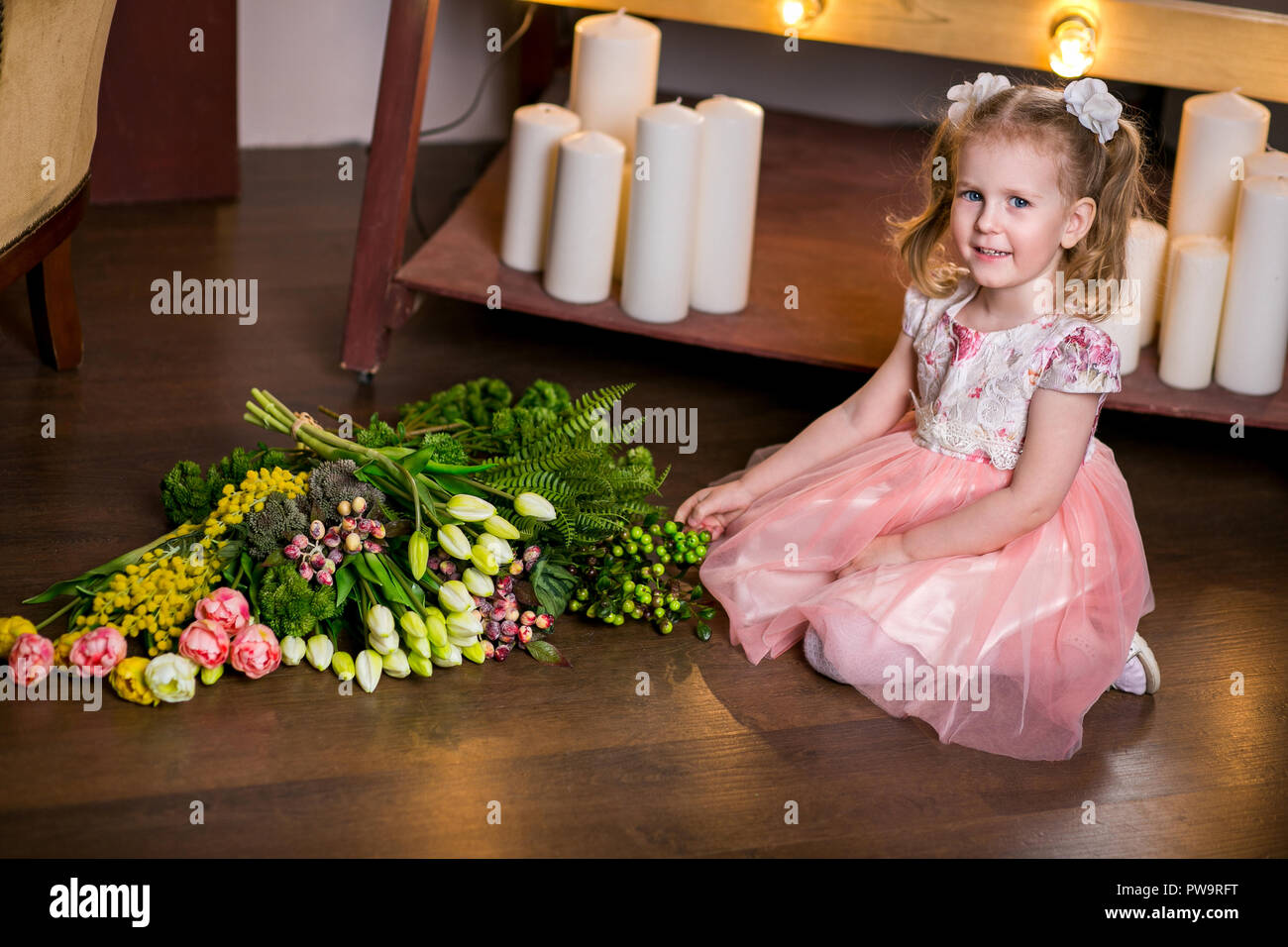 blue-eyed cute girl in a pink dress sits on the floor next to a bouquet of tulips, mimosa, berries and greenery Stock Photo