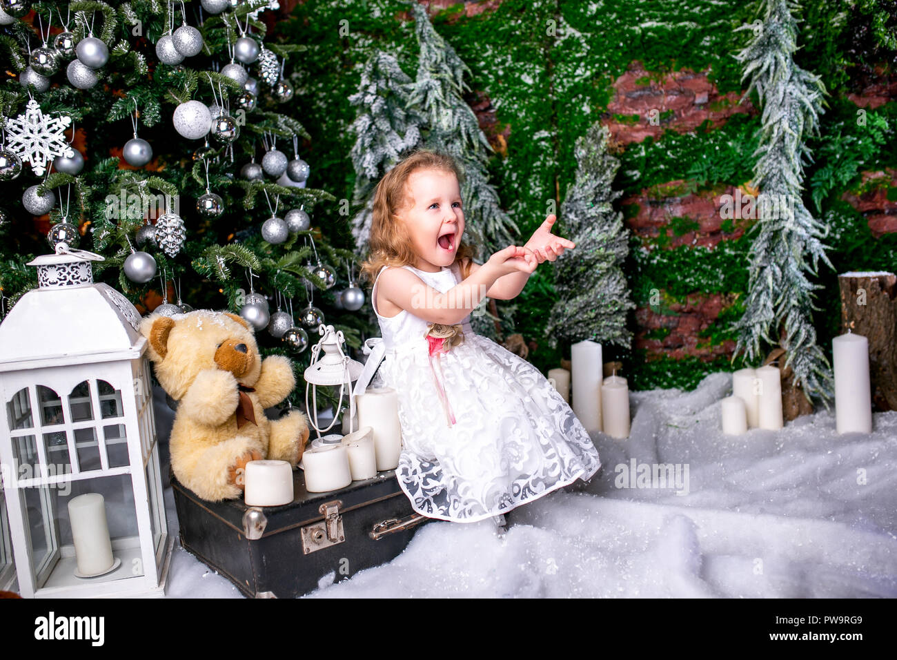 cute little girl in a white dress sitting near a Christmas tree on a suitcase next to the candles and a teddy bear, throws snow up and laughs Stock Photo