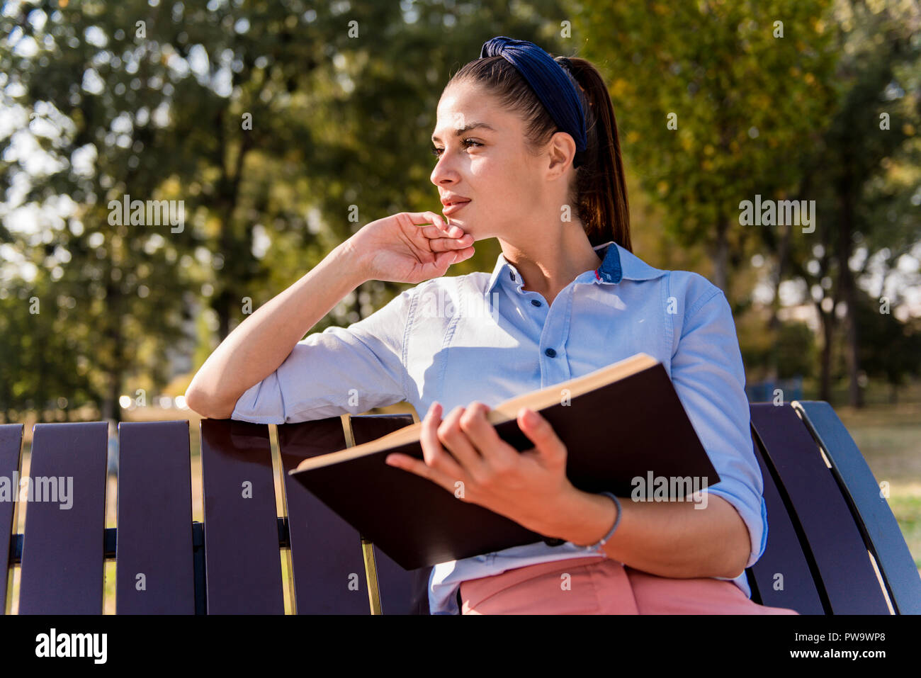 Cheerfull young woman holding open book and reading on bench in autumn ...