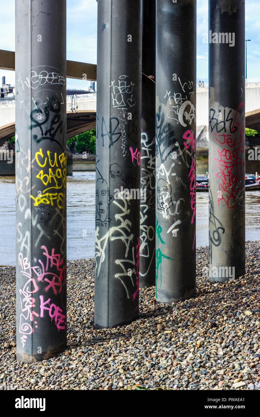 Graffiti-covered pillars beneath the Festival Pier at low tide on the Thames shore, London, UK Stock Photo