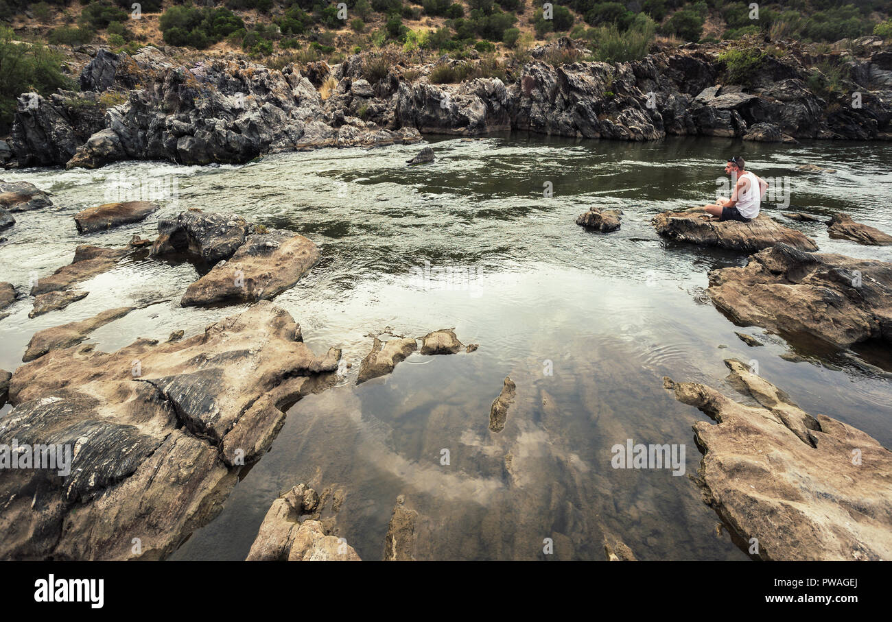 Young man in short white shirt sitting pensively on the rocks of the Guadiana river Stock Photo