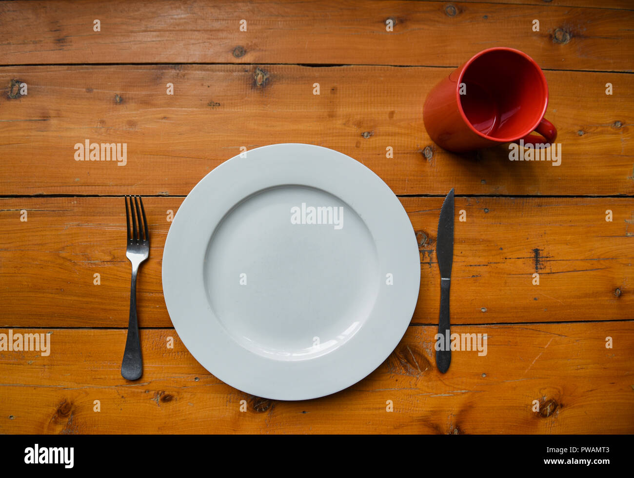 Top view of clean round white plate, silver fork and knife, red mug on a wooden table Stock Photo