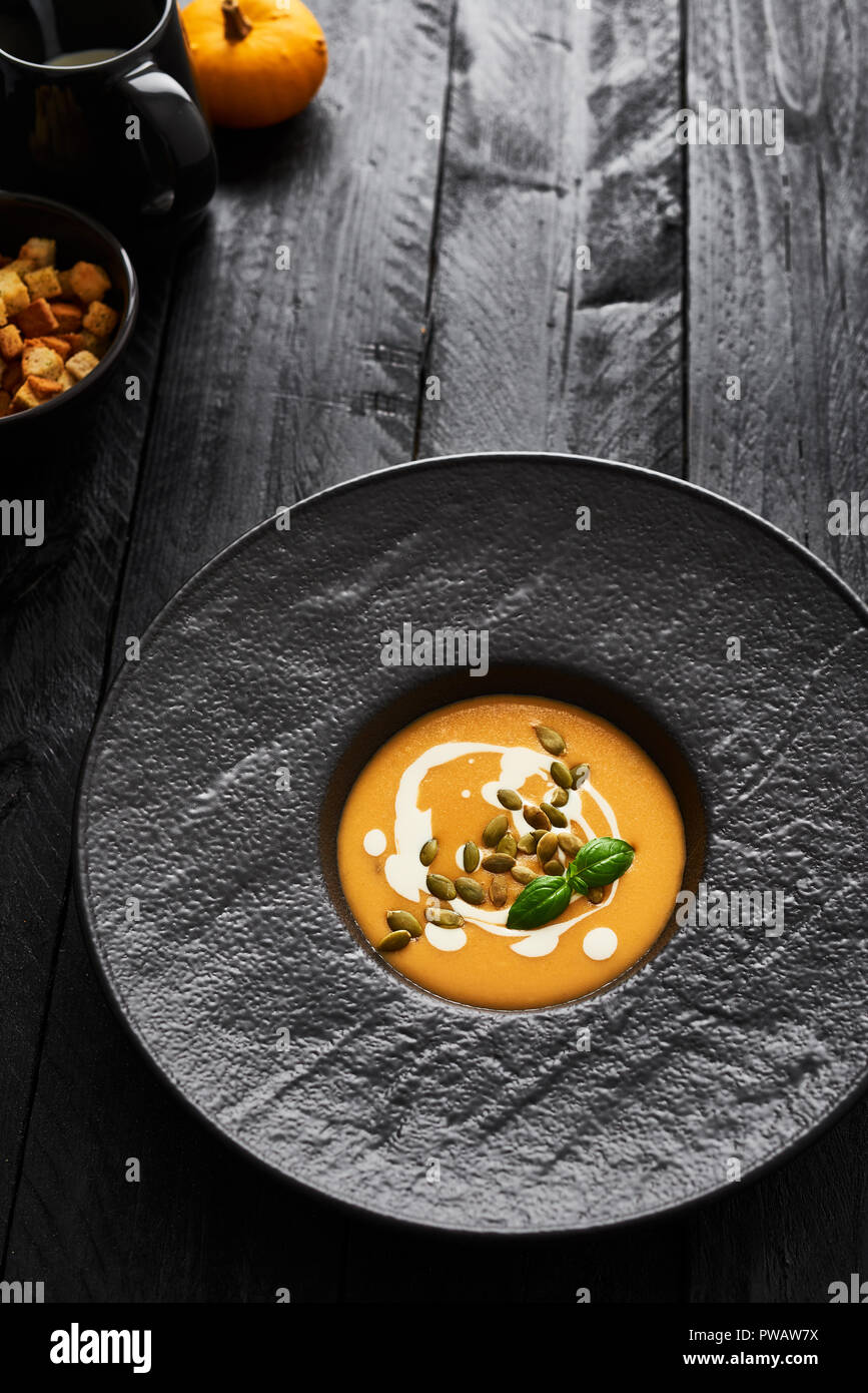 Pumpkin and carrot soup with pumpkin seeds, cream and basil in black modern plate on dark wooden table with copy space. Dark modern style. Top view. Stock Photo