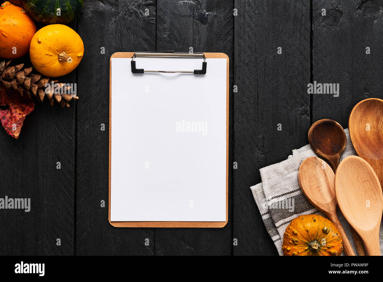 Autumn vegetables cooking preparation. Pumpkins, wooden spoons and blank paper on cardboard clipboard for menu or recipes on black wooden background.  Stock Photo