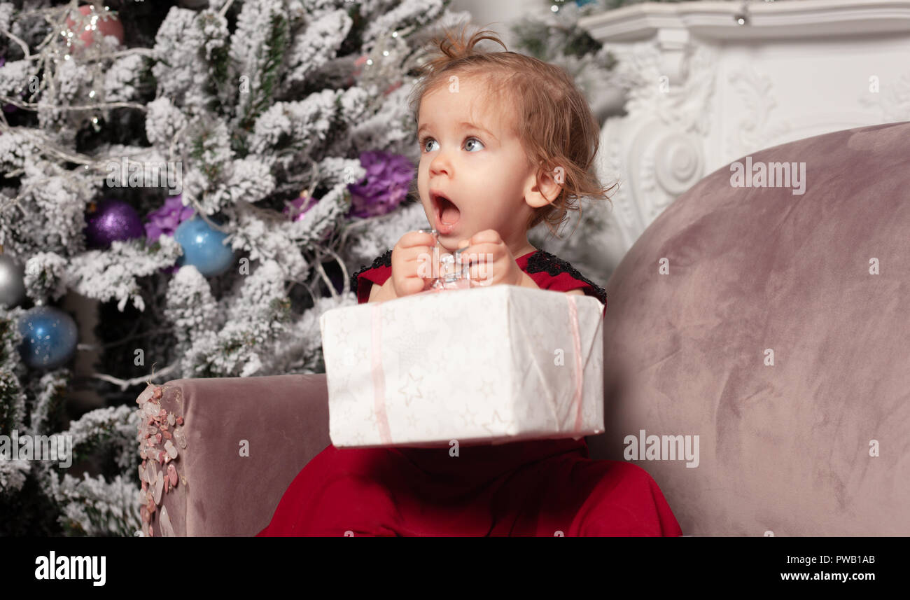 A beautiful cute little girl dressed in an elegant evening red dress sits on the couch and opens a New Year's gift. Against the background is a Christ Stock Photo