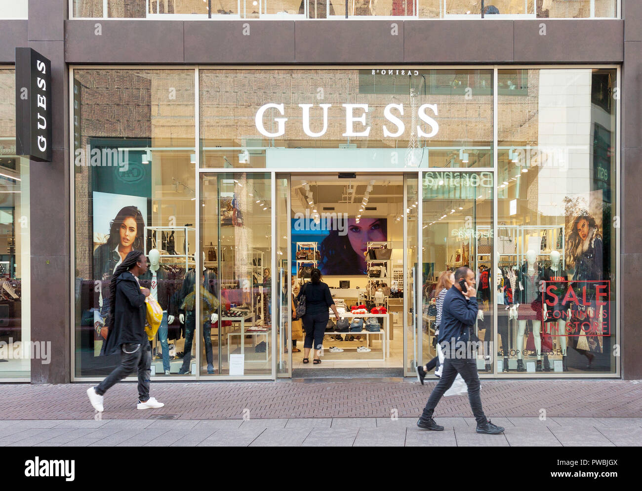 View of Guess luxury fashion house store entrance with brand signage and people passing by in The Hague, Net Stock Photo