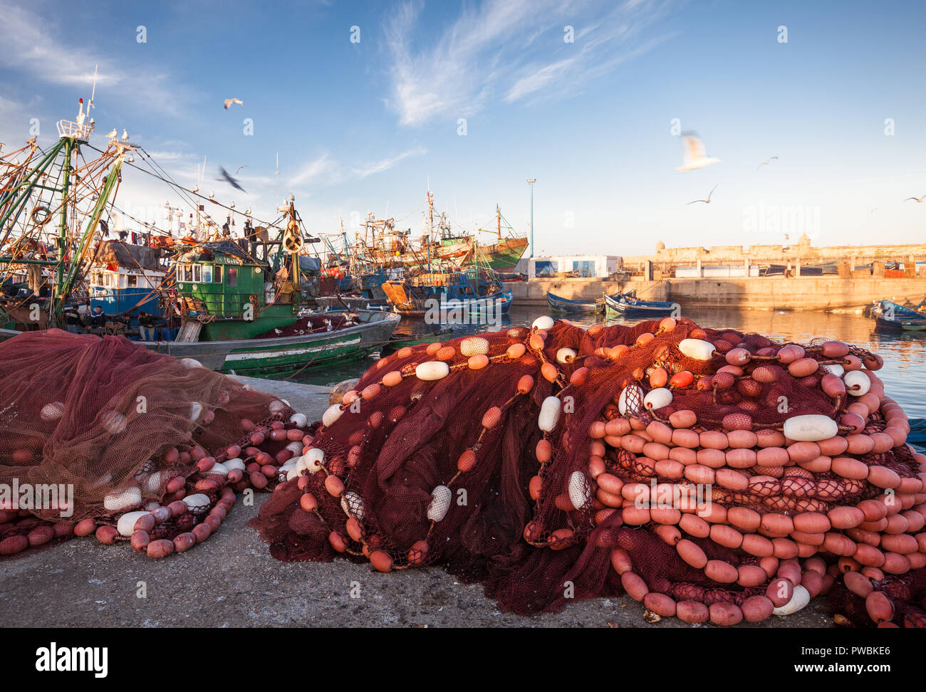 Morocco Essaouira harbor scene in daylight. Mounds of red fish netting and floats in the foreground, colorful painted fishing boats in the backtround. Stock Photo