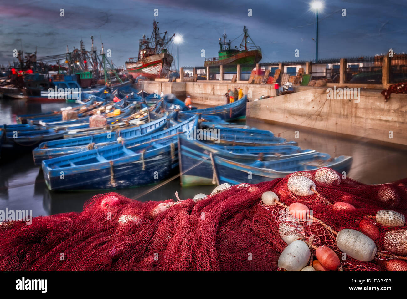 Morocco Essaouira harbor at night. Selective focus on the foreground red fish netting and floats. Background motion blur of blue boats and seagulls. Stock Photo