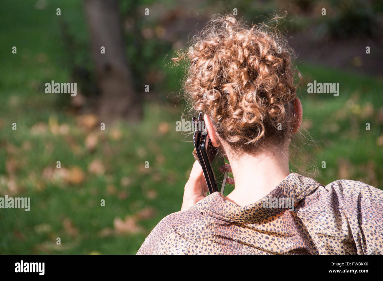 A red headed woman sitting alone on a park bench making a phone call on her mobile phone in London, UK Stock Photo