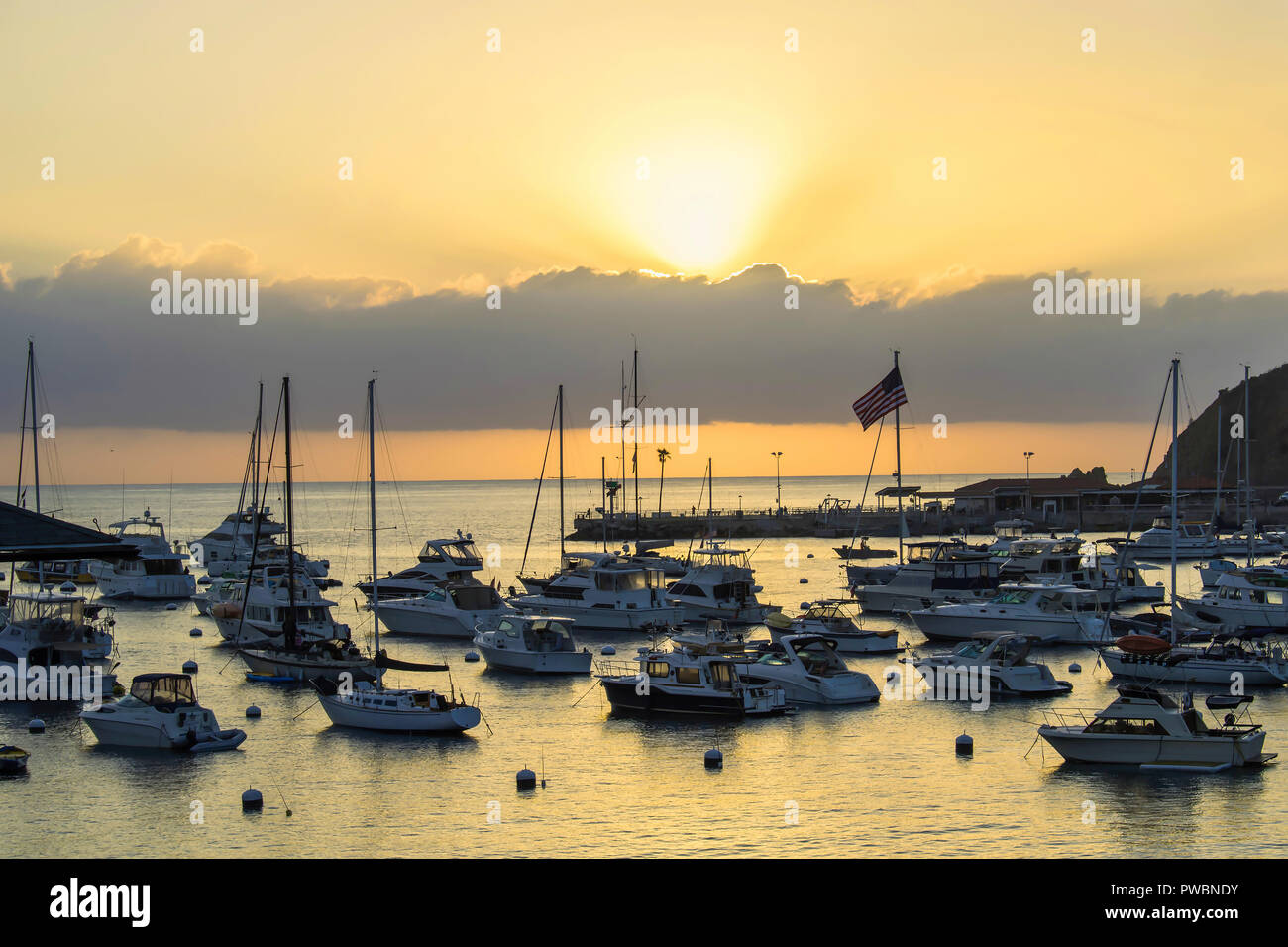 Daybreak with sun rays over harbor full of boats with orange light reflecting on ocean surface and American flag on horizon Stock Photo