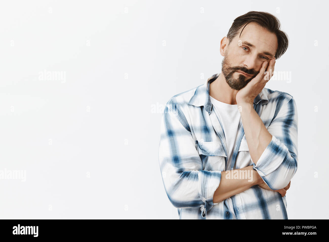So boring. Indifferent cute adult man with beard and dark hair, leaning face on palm and gazing with careless tired expression at camera, fed up of listening meeting for hours, bored and exhausted Stock Photo