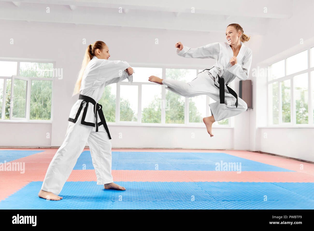 Two young women wearing in white kimono and black belt training karate martial arts against big window. One strong female fighter jumping and performing kick to other girl who making block of strike. Stock Photo