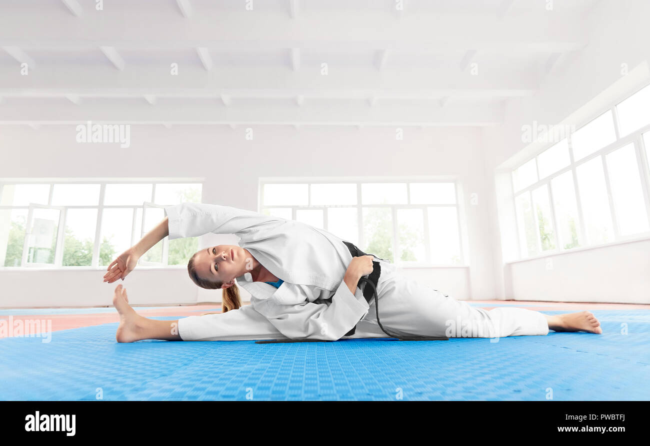 Young active girl wearing in white kimono with black belt, doing exercise on flexibility. Seriously sporty woman limbering up on floor before training in light gym. Healthy lifestyle concept. Stock Photo