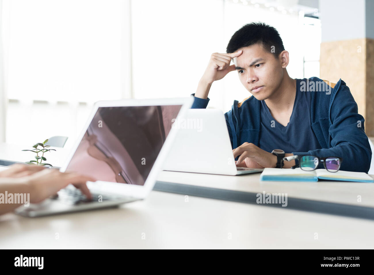Serious concentrated young Asian programmer focused on work sitting at table and installing setup for laptop in open space Stock Photo