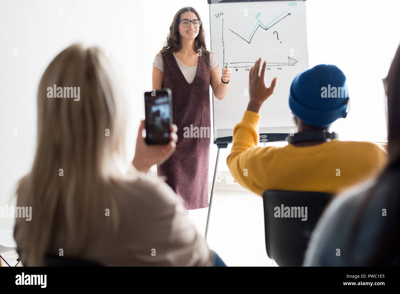 Cheerful confident female sales manager in glasses standing at whiteboard with graph drawing and holding seminar for students, she answering questions Stock Photo