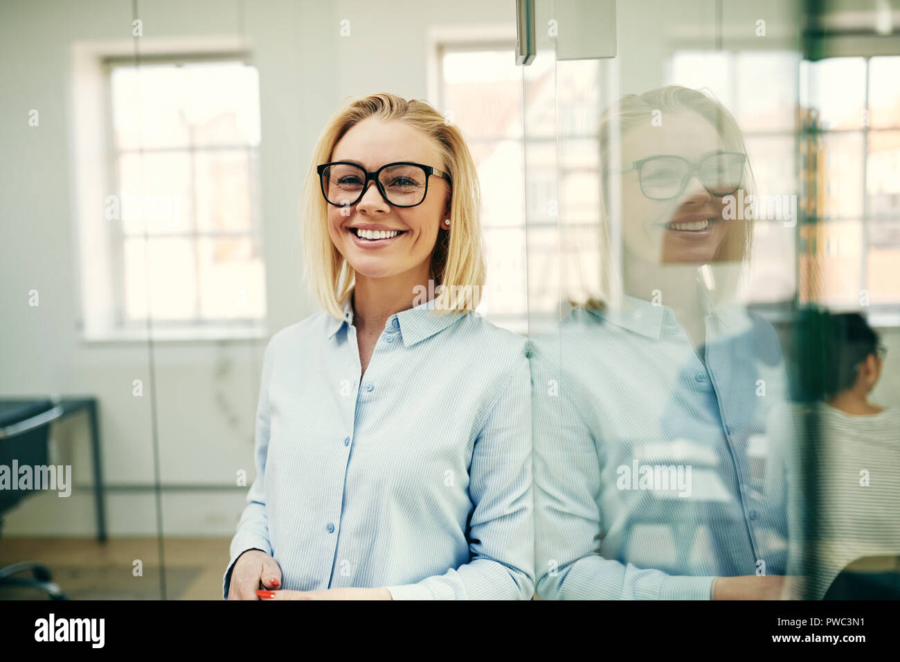 Young businesswoman smiling confidently while leaning against a glass wall in an office with colleagues in the background Stock Photo