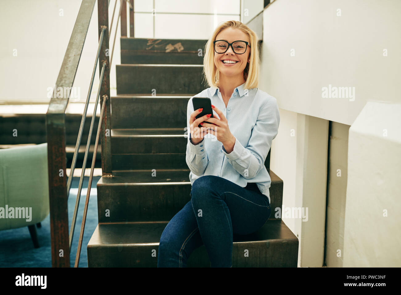 Young businesswoman wearing glasses and laughing at a funny a text message on her cellphone while sitting on stairs in a office Stock Photo