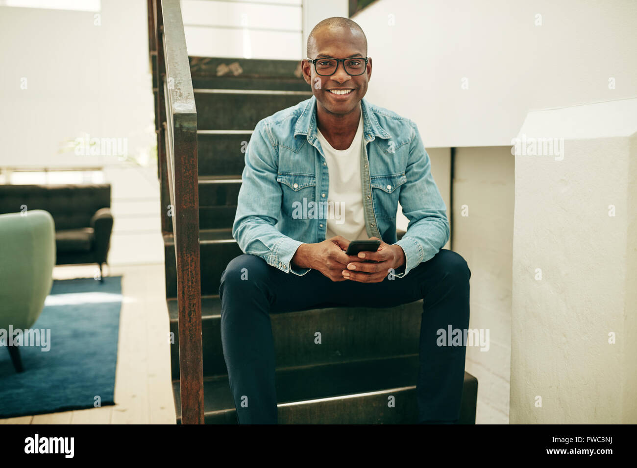 Smiling young African businessman wearing glasses sitting on stairs in an office reading text messages on a cellphone Stock Photo