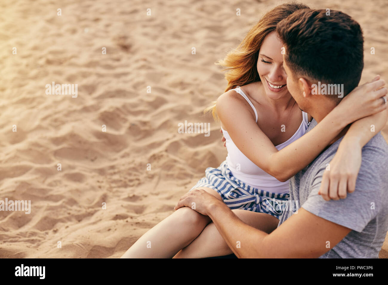Romantic young couple sitting arm in arm together on a sandy beach watching the sunset Stock Photo