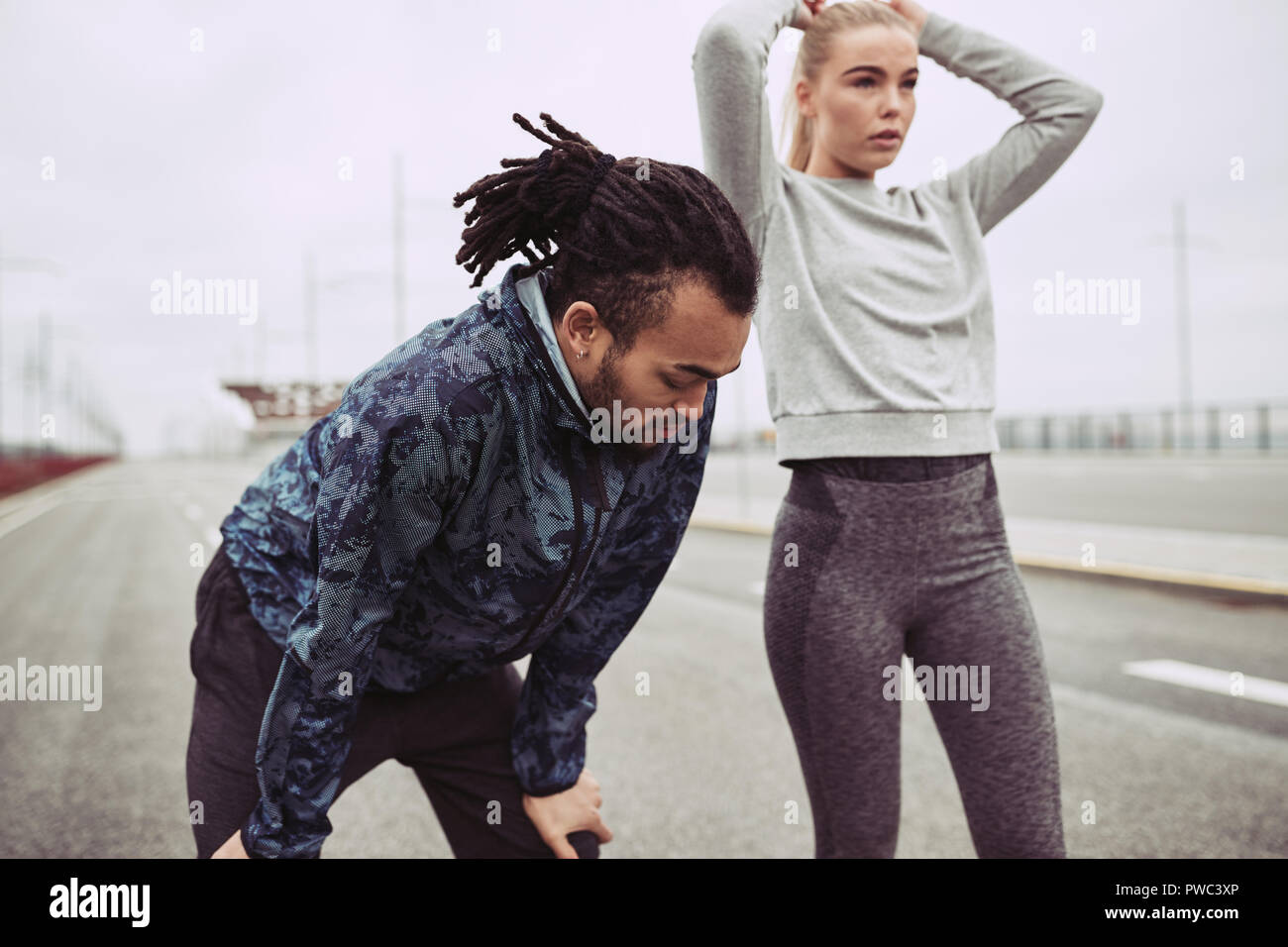 Diverse young couple catching their breath while taking a break from a run together on an overcast day Stock Photo
