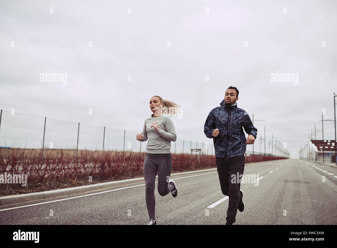 Diverse young couple in sportswear running together along a road in the country on an overcast day Stock Photo