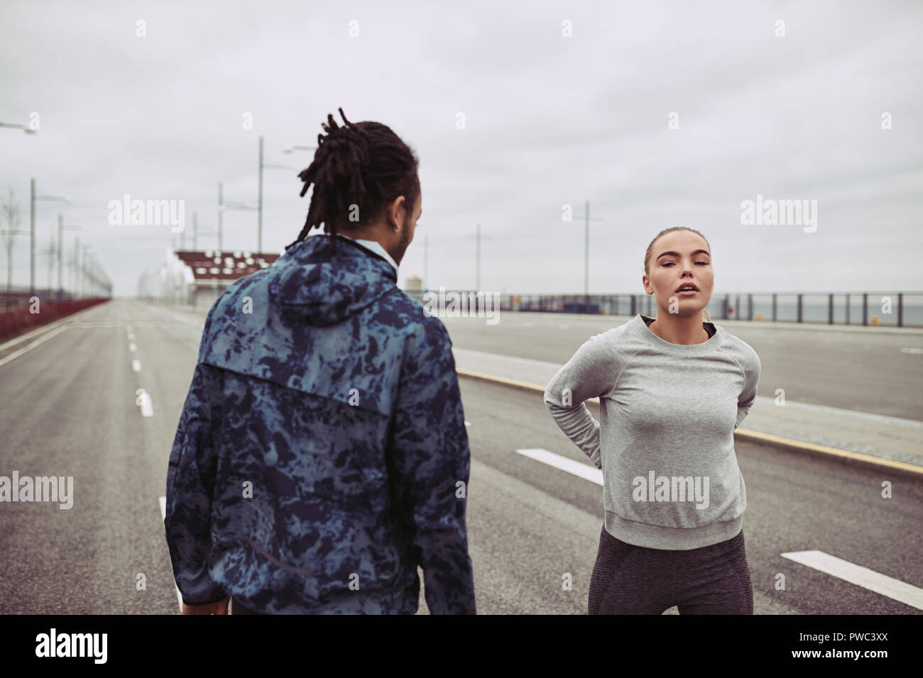Diverse young couple catching their breath and stretching while out for a run on an overcast day Stock Photo