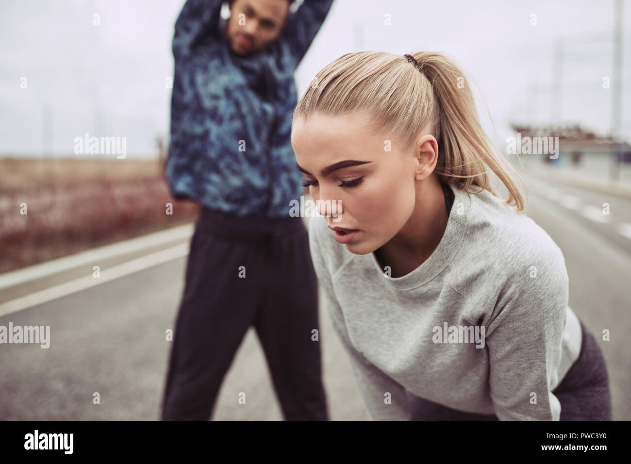 Young woman catching her breath with her boyfriend stretching in the background while out for a run on a country road together on an overcast day Stock Photo