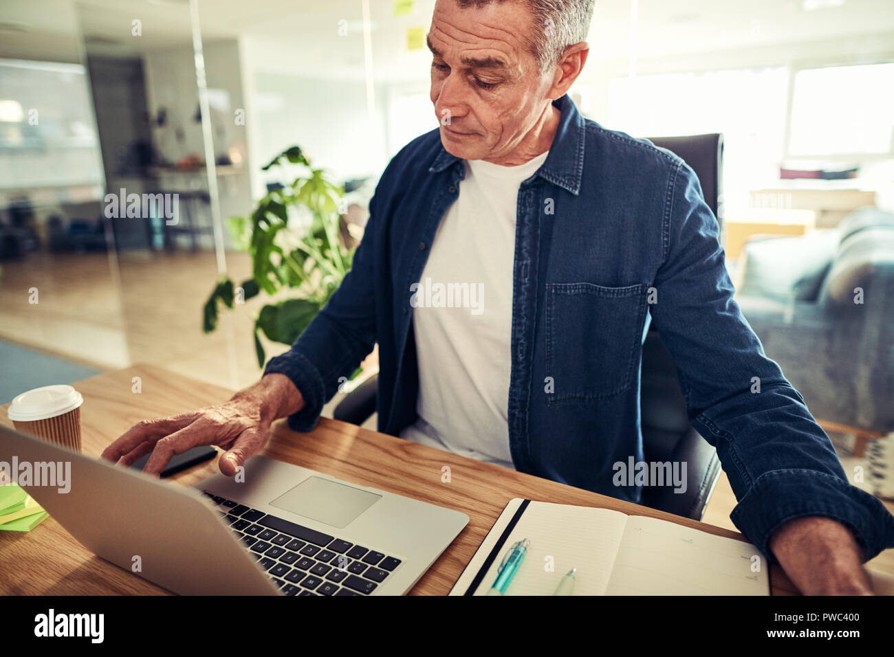 Casually dressed mature businessman sitting at his desk in an office working on a laptop and going over his schedule Stock Photo