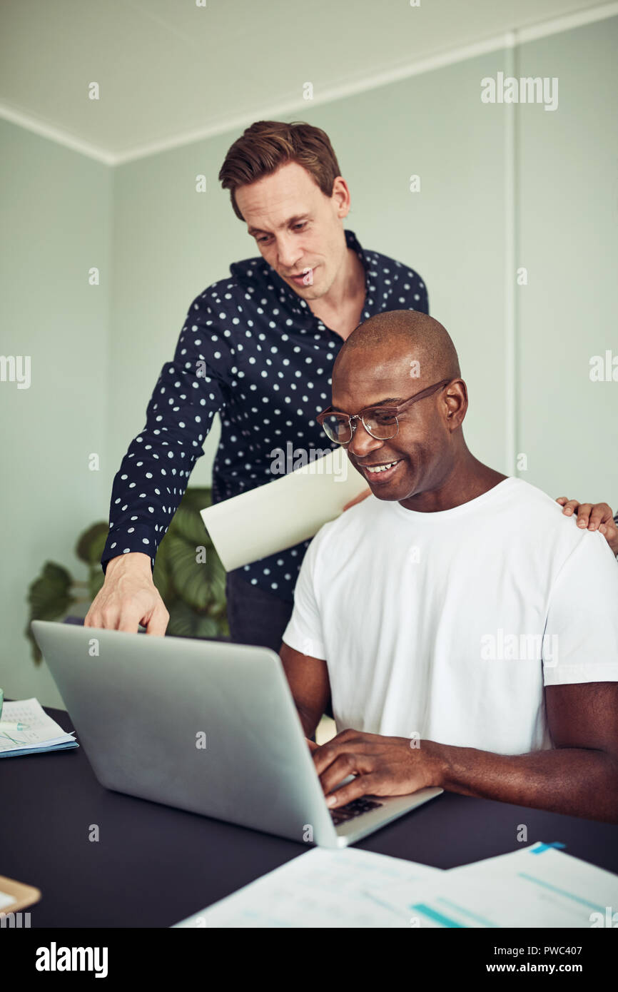 Smiling African businessman sitting at a desk in an office using a laptop and talking with a colleague standing behind him Stock Photo