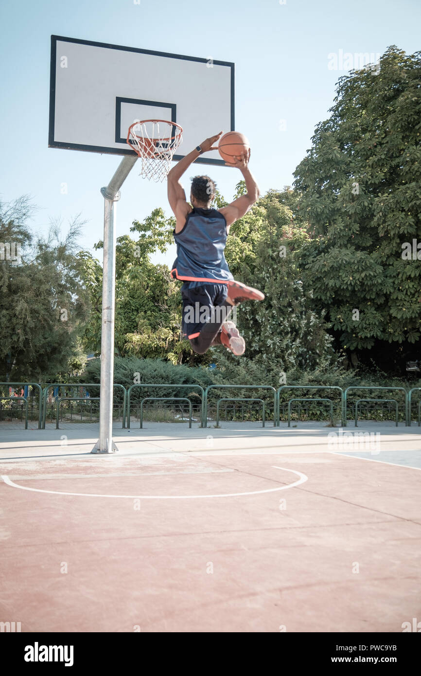 One afroamerican young man is playing basketball in a park in Madrid during summer at midday. He is doing an awesome jump to make a dunk and put the b Stock Photo