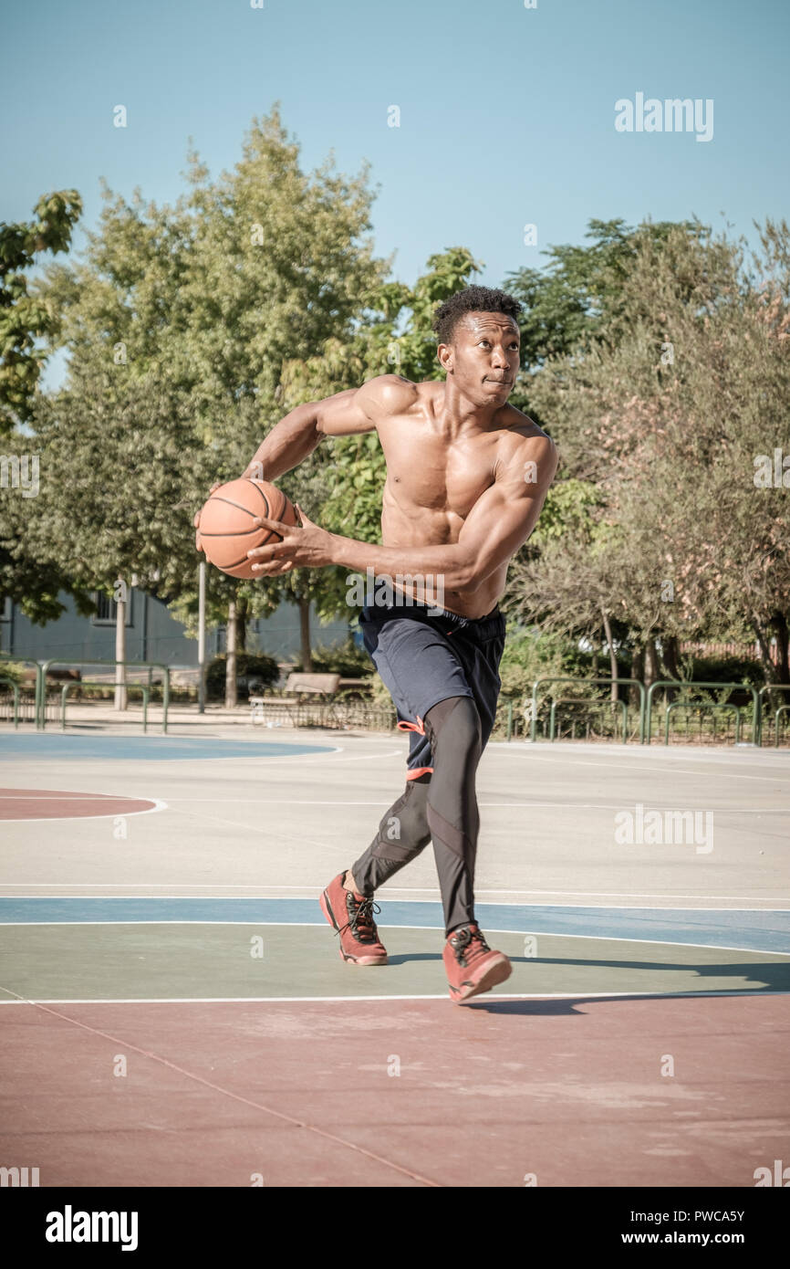 One afroamerican young man without tshirt is playing basketball in a park in Madrid during summer at midday. He is doing an awesome jump to make a dun Stock Photo