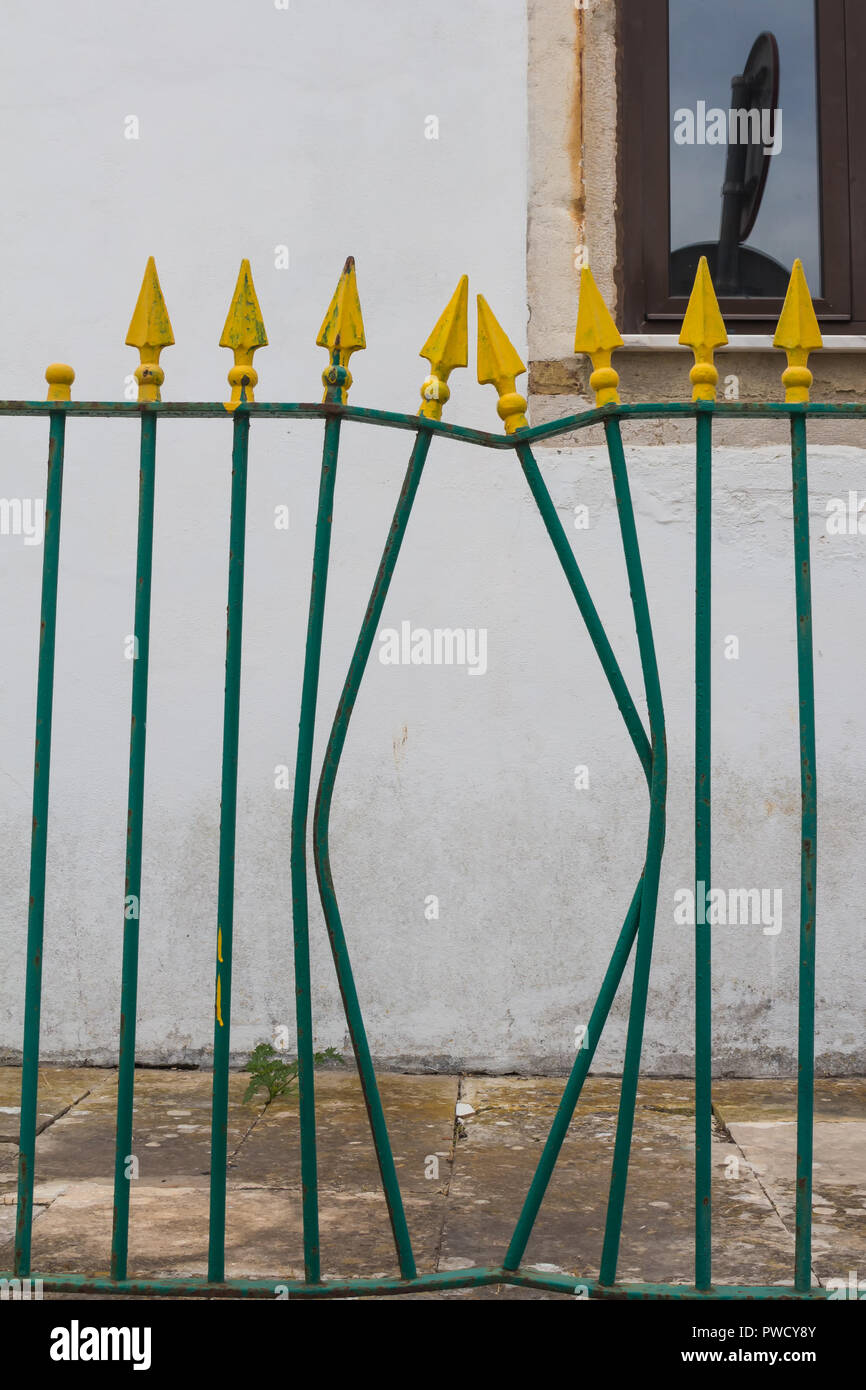 Bars of the fence painted green with yellow spokes. Part of the fence deformed. White wall and a window with a reflection in the background. Lagos, Al Stock Photo