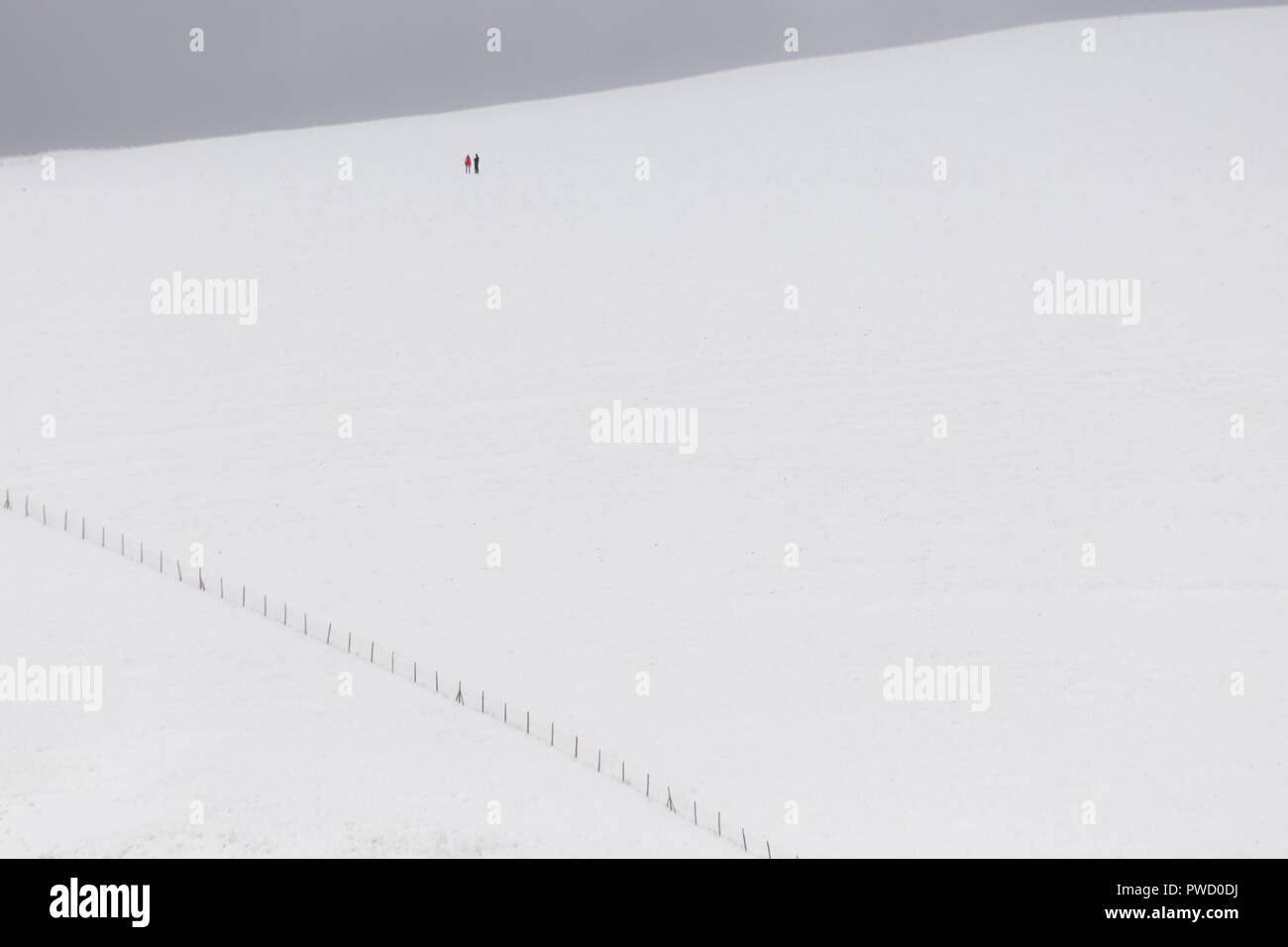 A very minimalistic view of two distant people over a mountain covered by snow, near a fence Stock Photo